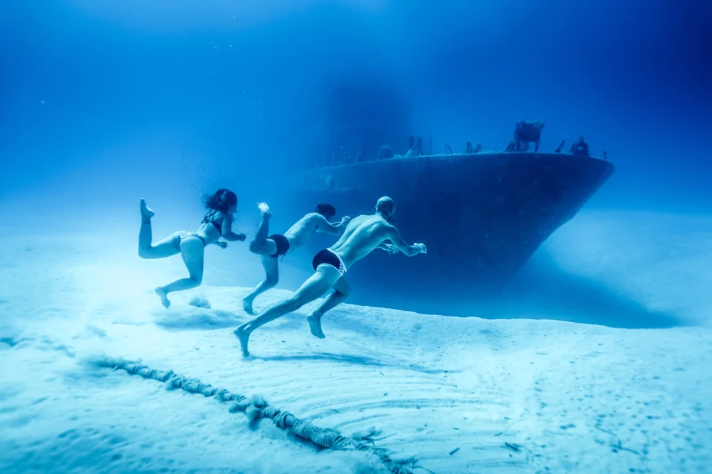 Three freedivers run on sand towards ship's bow
