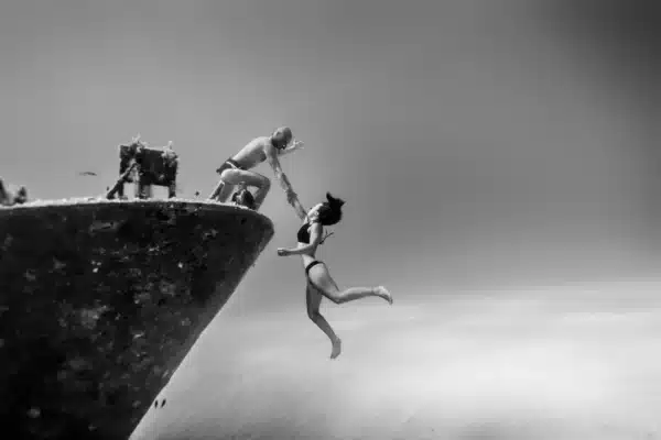 A freediver grabs another freedivers arm on the bow of a ship